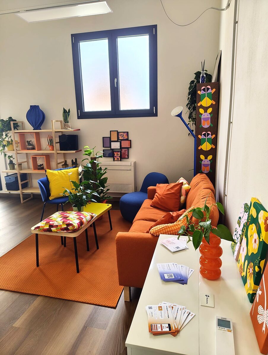L'angolo lounge all'ingresso: divano arancione, libreria con le piante e il tavolo con i materiali